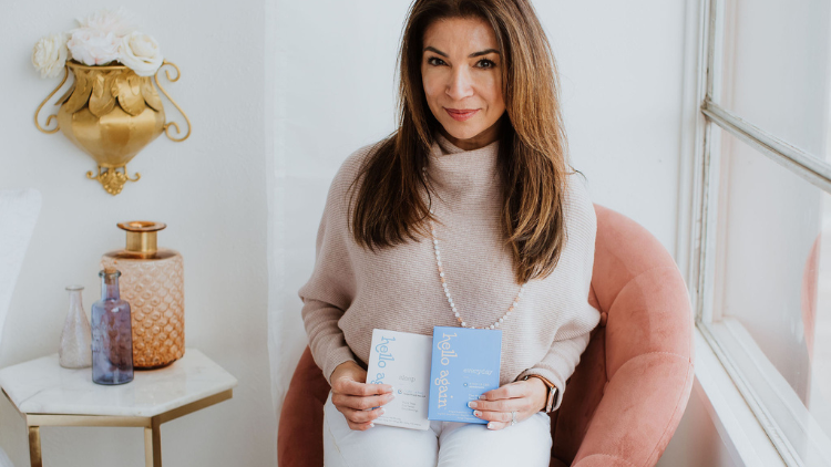 A middle-aged woman holds Sleep and Menopause Hello Again products while sitting in a velvet pink chair in the corner of a bedroom.