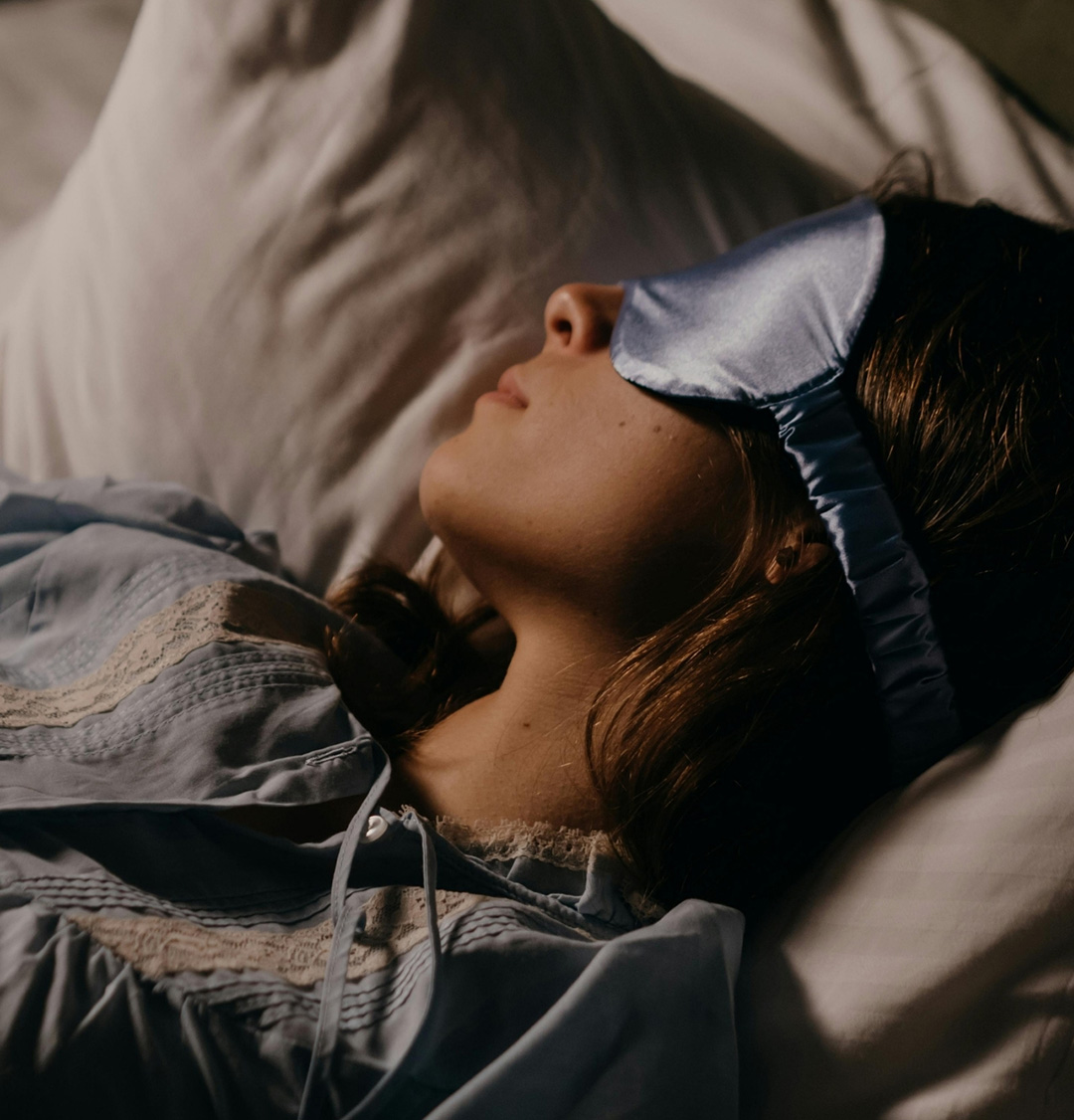A woman sleeps peacefully in a dark room, a blue silk sleep mask resting gently over her eyes.