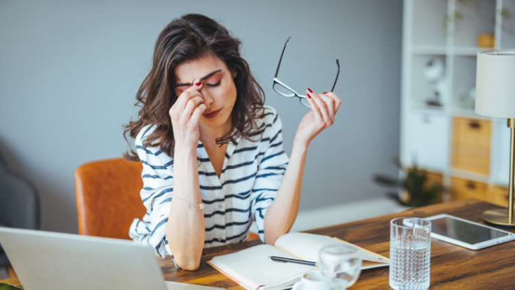 A stressed woman sits at a desk, elbows resting on the surface, pinching the bridge of her nose while holding her glasses in her left hand.