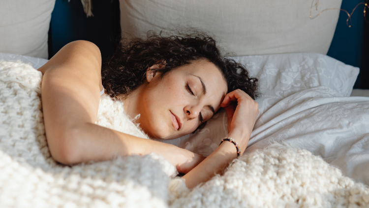 A woman sleeps peacefully on white bedding, wrapped in a textured blanket.