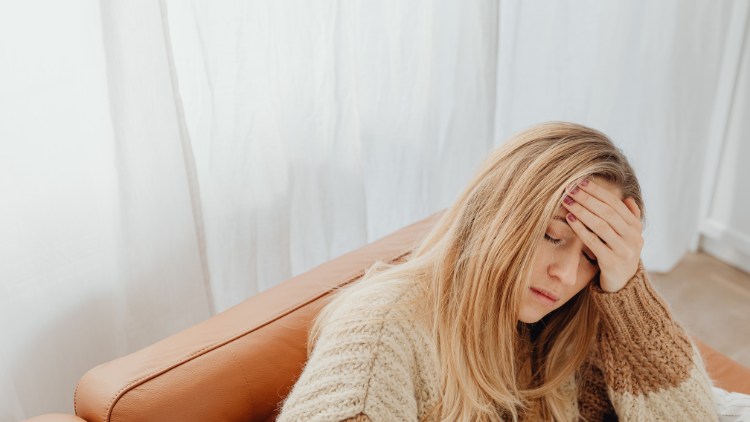 Blonde woman sitting on a couch with her hand on her forehead, appearing tired and uncomfortable.