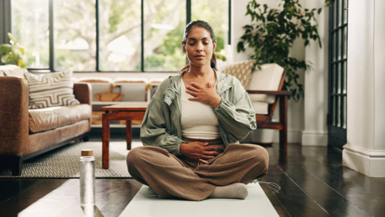 A woman sitting cross-legged on a yoga mat in a sunlit living room, practicing mindful breathing with one hand on her chest and the other on her abdomen.