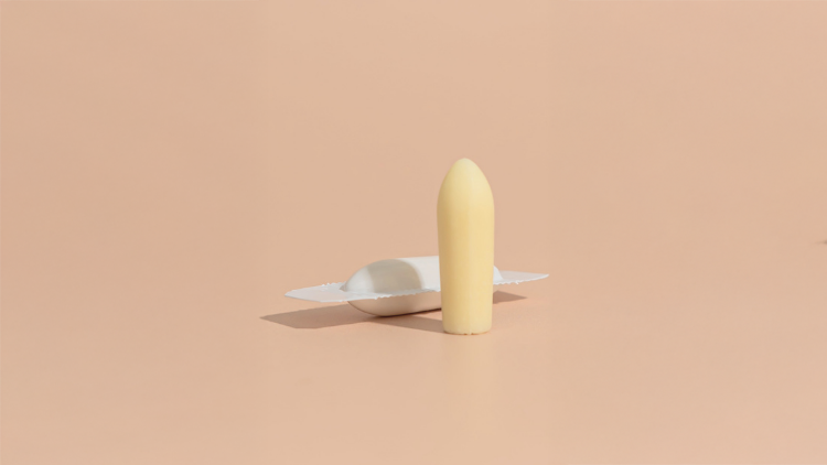 A Hello Again vaginal suppository standing upright beside its open wrapper against a soft peach background, highlighting plant-based natural wellness practices.