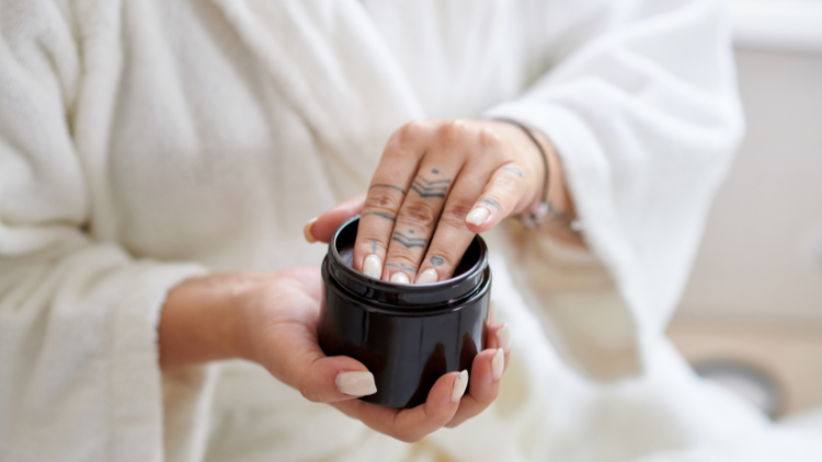 Close-up of a woman in a white robe applying skincare from a black jar, highlighting clean beauty and skincare as part of a natural wellness routine.